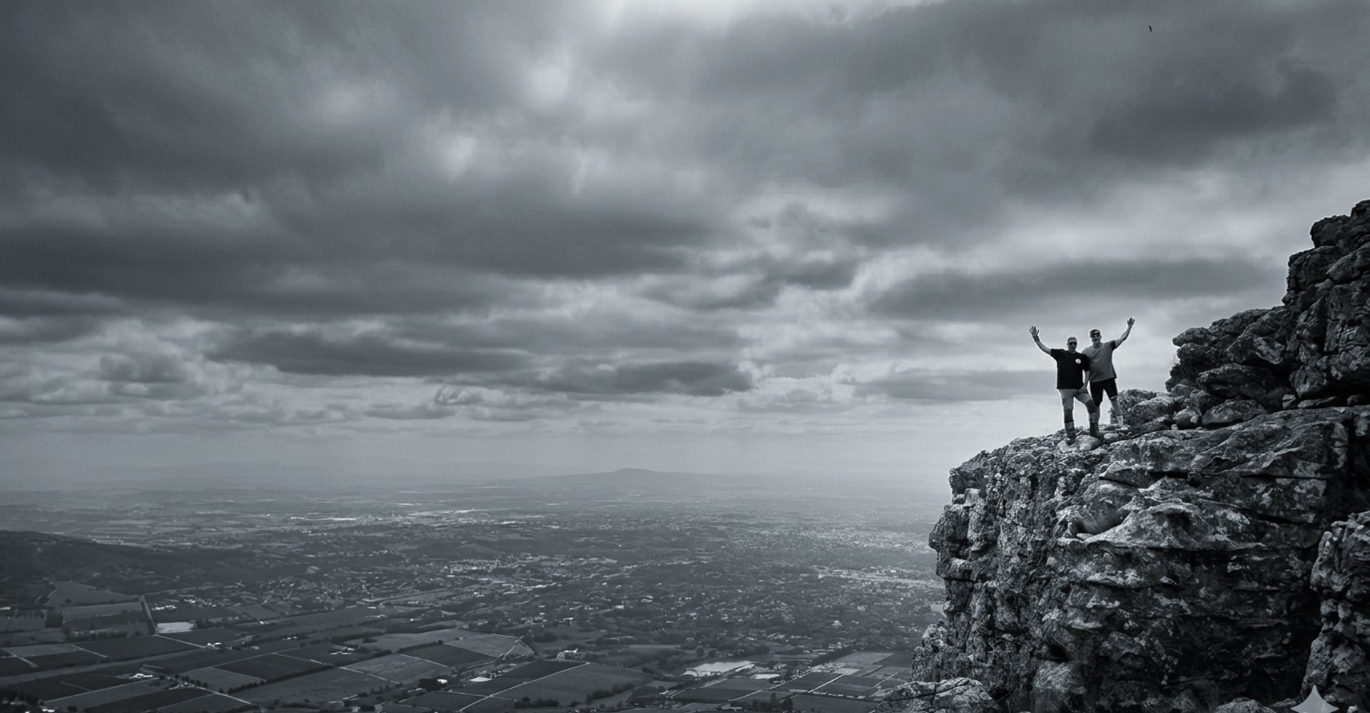 Two figures stand victorious on a mountain cliff edge overlooking a vast valley under dramatic clouds